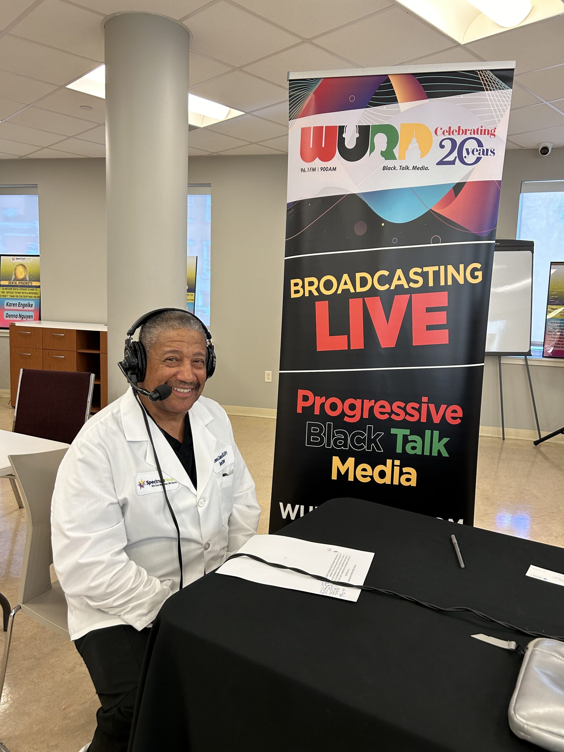 A guest in a white coat wears headphones at the radio table