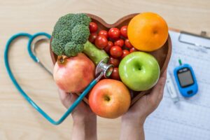 A heart-shaped bowl filled with apples, an orange, broccoli, tomatoes, and onion with a stethoscope and blood sugar monitor in the background