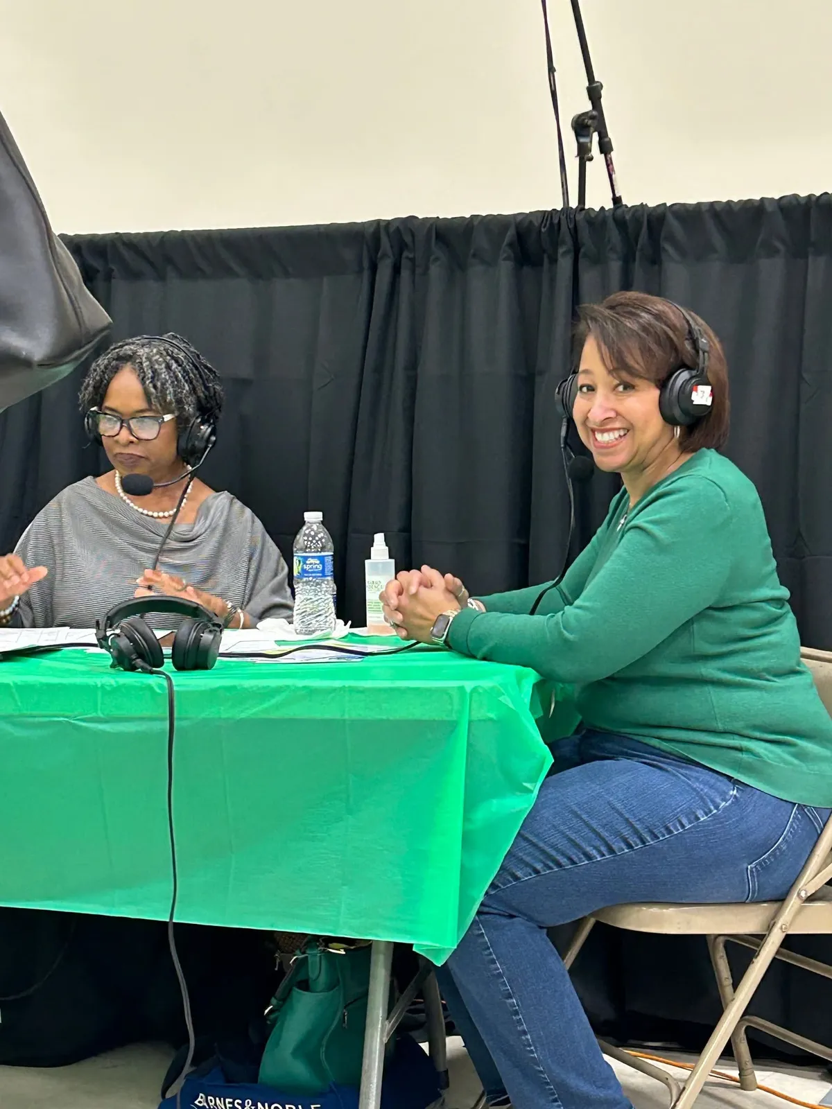 Two women wearing headphones smile during a live radio talk at a green table.