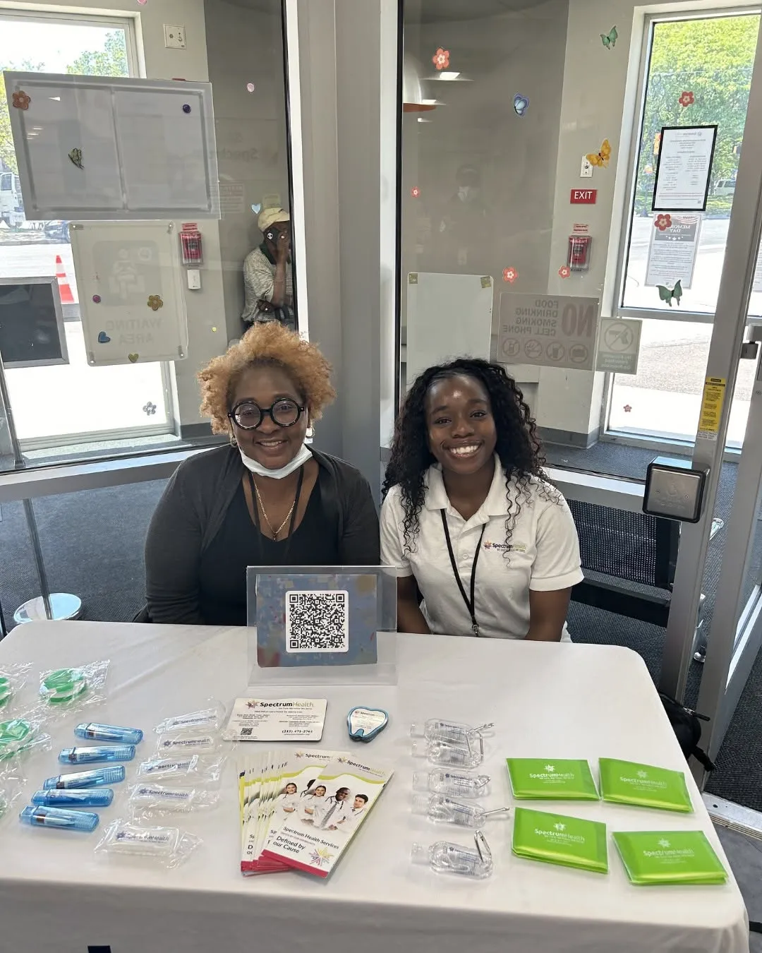 Two smiling Spectrum staff members sit at a table with health flyers and giveaways.
