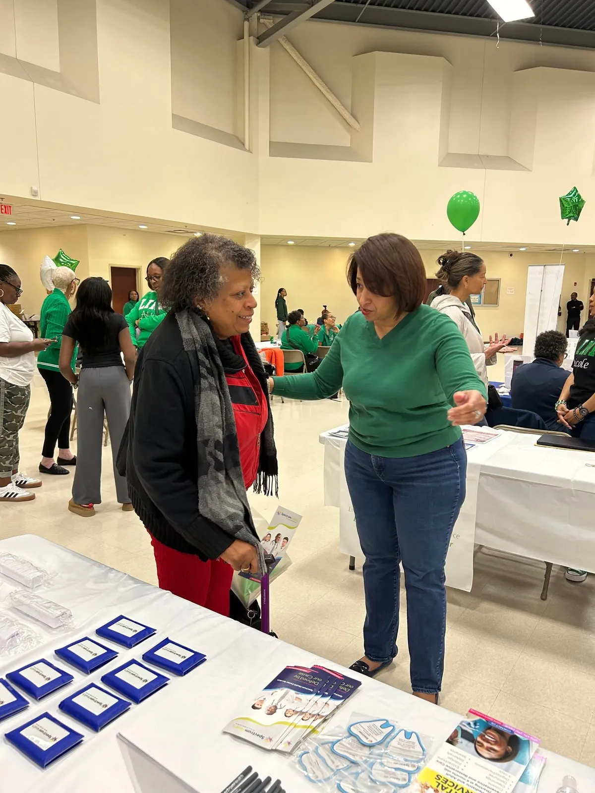 Two women chat at a table full of Spectrum giveaways and health flyers.
